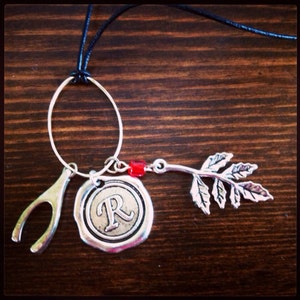 May include: A silver-toned necklace with a black cord. The necklace features a wishbone charm, a circular pendant with the letter "R", a red bead, and a leaf-shaped charm. The charms are suspended from a silver oval.