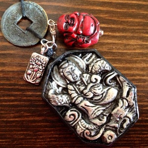 May include: A collection of Asian-inspired pendants and charms. Includes a weathered coin, a red carved figure, a silver floral charm, and a large, detailed pendant with a figure in relief. The items are displayed on a wooden surface.