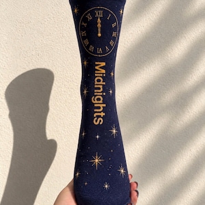 Puede incluir: Un calcetín azul marino con una esfera de reloj dorada con números romanos marcando la medianoche. La palabra Midnights está impresa verticalmente en letras doradas debajo del reloj, rodeada de varias estrellas doradas en la tela.