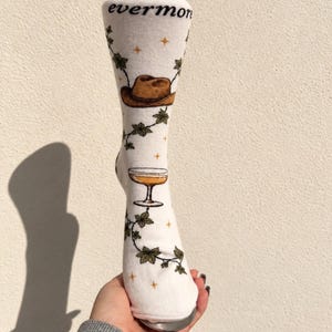 Peut inclure: Une chaussette montante blanche avec le mot evermore imprimé en haut. Le motif comprend un chapeau de cowboy marron, une coupe à cocktail sur pied et des vignes de lierre vertes s'enroulant autour, agrémentées de petites étoiles orange.