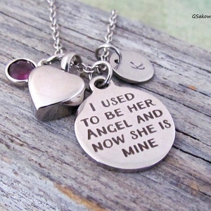 May include: A silver necklace with multiple charms. Charms include a heart-shaped pendant, a round pendant with the inscription "I USED TO BE HER ANGEL AND NOW SHE IS MINE", a small disc with the letter "K", and a purple gemstone.