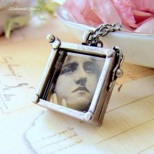 May include: A silver-toned pendant necklace featuring a square frame with a vintage portrait inside. The frame has decorative corner accents and is suspended from a delicate chain. The image is a black and white portrait of a man.