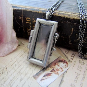 May include: A rectangular, silver-toned locket pendant with a hinged opening, suspended from a matching chain. The locket is displayed on a vintage paper background with a small image of a woman.