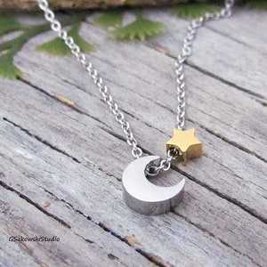May include: A silver-toned necklace featuring a crescent moon pendant and a gold-colored star charm. The chain is delicate, and the pendants are attached with small rings. The necklace is displayed on a weathered wooden surface.