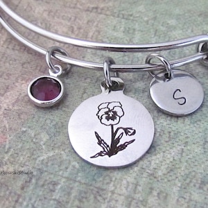 May include: Silver-toned adjustable bangle bracelet with three charms. One charm is a round, dark purple gemstone. Another is a circular charm with a flower engraving. The third charm is a small disc with the letter "S".