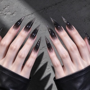 May include: Long, stiletto-shaped press-on nails featuring a translucent black ombre gradient that fades into a clear base at the cuticle. The nails have a sharp, pointed tip and a glossy finish, worn on fingers with black leather sleeves.