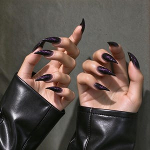 May include: Stiletto-shaped press-on nails featuring a dark black base with a purple lightning bolt pattern. The nails have a glossy finish and are displayed on fingers wearing black leather sleeves, showcasing a sharp, pointed tip design.
