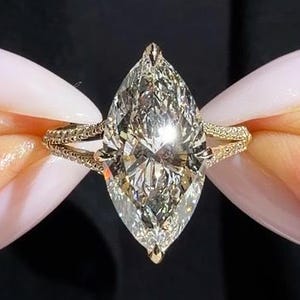 May include: A marquise-cut diamond ring featuring a large, elongated center stone set in a gold band. The band has a split-shank design adorned with small pavé-set diamonds along the sides, creating a brilliant, reflective appearance.