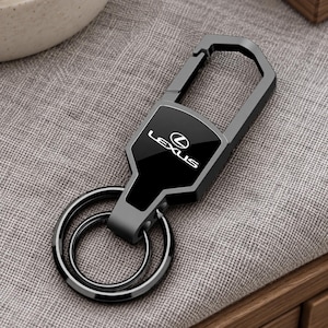 Pode incluir: Um porta-chaves de metal cinzento-escuro com um mosquetão no topo e dois anéis circulares na base. A placa retangular central tem um acabamento preto brilhante com o logótipo e o texto Lexus impressos a branco no centro da peça.