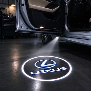 Puede incluir: Una puerta de coche plateada está abierta, revelando un panel interior beige. Una luz montada en la parte inferior de la puerta proyecta un logotipo de Lexus circular, blanco y azul, sobre el suelo, iluminándolo con un gráfico nítido.