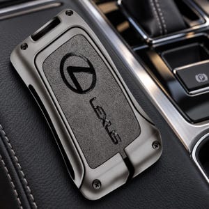 Puede incluir: Una funda para llave de coche Lexus en color gris metálico con un panel central negro texturizado que presenta el logotipo y el nombre de la marca Lexus en relieve. La carcasa protectora rectangular se fija con pequeños tornillos visibles.