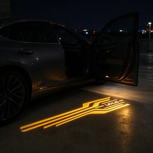Puede incluir: Un sedán gris oscuro con la puerta del pasajero abierta, que proyecta una luz LED amarilla brillante en el suelo. La luz muestra un patrón geométrico de líneas paralelas y la palabra LEXUS en un estilo de fuente sans-serif limpio.