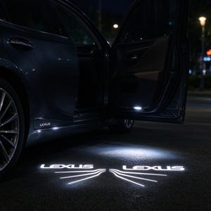 Puede incluir: Un vehículo Lexus de color oscuro con la puerta abierta, que proyecta una luz LED blanca brillante sobre el suelo. La proyección muestra la palabra LEXUS en una fuente estilizada con líneas radiantes, iluminando la superficie oscura.