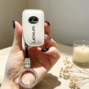 Puede incluir: Una funda de cuero blanco para llave con el logotipo y el texto de Lexus en negro. La funda está unida a una correa negra conectada a un anillo metálico circular incrustado con pequeños cristales transparentes, un accesorio funcional.