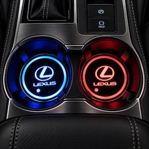 Peut inclure: Deux inserts de porte-gobelets circulaires pour voiture avec le logo et le nom de la marque Lexus illuminés en bleu et rouge vifs. Les inserts sont placés dans un panneau de console centrale en métal brossé avec des boutons de frein électronique.
