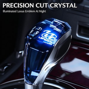 Puede incluir: Un pomo de palanca de cambios de cristal azul tallado con precisión que muestra el emblema de Lexus y el texto PRECISION CUT CRYSTAL e Illuminated Lexus Emblem At Night. El pomo tiene un cuerpo azul translúcido con detalles laterales plateados.