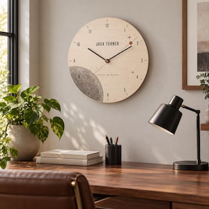 Puede incluir: Un reloj de pared circular de madera clara con números negros del 1 al 12, manecillas negras y un gráfico de luna gris texturizado en la parte inferior izquierda. El texto JACK TURNER y BEGIN ANYWHERE está impreso en negro en la esfera.