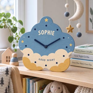 May include: A cloud-shaped wall clock in blue and yellow with dark blue clock hands. The clock face features the name SOPHIE in light yellow letters, small yellow stars, dark blue dots, and the text GOOD NIGHT printed in dark blue.