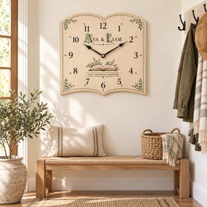 Peut inclure: Une horloge murale en bois clair en forme de livre ouvert. Le cadran présente des chiffres noirs, des aiguilles noires, et le texte "Ava & Liam" au-dessus d'une illustration de livre ouvert avec les mots "Our Favorite Chapter" en dessous.