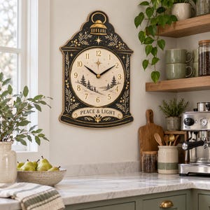 May include: A wall clock with a black frame and a cream-colored face featuring a winter forest illustration, black hands, and the text PEACE & LIGHT. The clock has a decorative top with a lantern icon and gold-colored botanical accents.