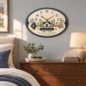 May include: An oval wall clock with a cream face, dark blue border, and gold hands. The design features a house illustration, a large key, green foliage, and the text "THE BROWN HOME" and "KEYS TO NEW BEGINNINGS" with mixed numerals.