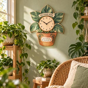 May include: A wall clock shaped like a potted plant with green variegated leaves. The clock face features black numbers and hands. A banner at the top reads THE EVANS HOME, and a label on the terracotta-colored pot reads ROOTED HERE 2026.