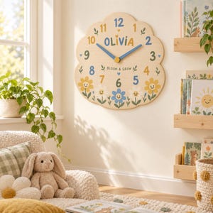 May include: A scalloped, light-wood wall clock featuring the name OLIVIA in blue and yellow text. The clock face displays colorful numbers, blue hands, and a floral design with smiling faces, along with the text BLOOM & GROW in the center.