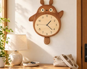 Puede incluir: Un reloj de pared de madera marrón con forma de criatura de dibujos animados con dos orejas, una cara redonda y una esfera circular clara con números negros del uno al doce y dos manecillas negras que indican la hora actual.
