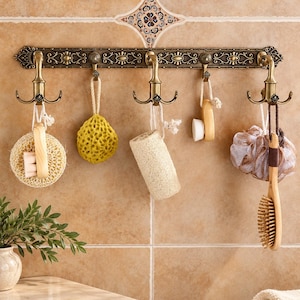 May include: A wall-mounted bronze-toned metal rack featuring five ornate hooks with intricate detailing. Hanging from the hooks are various bath accessories, including a round loofah, a yellow sponge, a cylindrical loofah, a small brush, and a hairbrush.