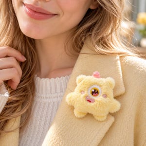 May include: A fuzzy, pale yellow plush monster brooch with a single embroidered eye, a small pink horn on top, and pink cheeks. The creature has a wide, smiling mouth with visible teeth and is attached to the lapel of a light yellow coat.