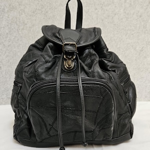 Könnte beinhalten: Ein schwarzer Rucksack aus mehreren zusammengenähten Lederstücken. Er verfügt über einen Tragegriff, eine Klappe mit einer bronzefarbenen Metallschnalle, zwei seitliche Reißverschlusstaschen und eine große vordere Reißverschlusstasche mit Lederbändern.