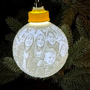 May include: A spherical white ornament featuring a 3D-printed relief of a family portrait on its surface. The ornament is topped with a bright yellow cap and a silver hanging loop, emitting a soft internal glow that highlights the faces.
