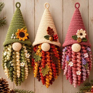 Puede incluir: Tres adornos colgantes de gnomos tejidos a ganchillo con gorros puntiagudos. El gnomo de la izquierda tiene un gorro verde con un girasol, el central un gorro crema con una bellota y hojas, y el de la derecha un gorro rosa con una flor blanca.