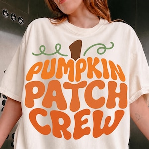 May include: A cream-coloured short-sleeve t-shirt featuring a large graphic print of a pumpkin shape with a brown stem and green vines. The text PUMPKIN PATCH CREW is printed in bold, wavy orange letters across the centre of the pumpkin design.