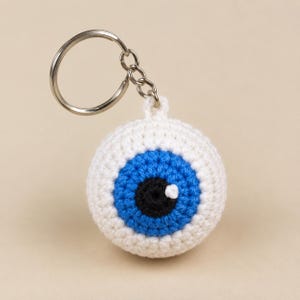 May include: A spherical crochet keychain shaped like an eyeball. The design features a white sclera, a bright blue iris, and a black pupil with a small white stitch detail representing a light reflection. It is attached to a silver metal ring.