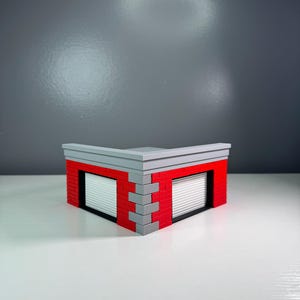 May include: A corner building model constructed from interlocking plastic bricks. The structure features red brick walls, gray corner detailing, a gray roof trim, and two white garage doors with horizontal slats set into black frames.