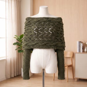 May include: An olive green off-the-shoulder sweater featuring a chunky cable knit pattern across the chest and shoulders, with long sleeves and a ribbed texture. The garment is displayed on a white mannequin torso against a neutral background.