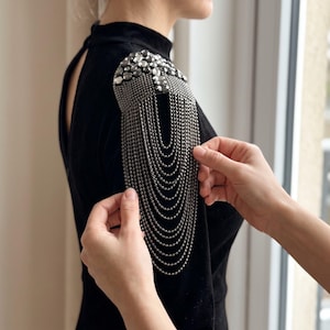 May include: A black long-sleeved top featuring a decorative shoulder epaulet. The epaulet is constructed from dark metallic beads and faceted crystals, with multiple cascading silver-toned ball chains hanging down from the shoulder piece.