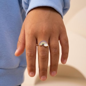 May include: A gold-toned metal ring featuring a small, colorful enamel rainbow charm with a white cloud at one end. The rainbow design includes bands of purple, blue, green, yellow, and pink, centered on a thin, polished metallic band.