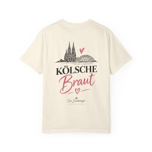 May include: An off-white short-sleeve t-shirt featuring a black graphic of the Cologne Cathedral and Hohenzollern Bridge with a pink heart. The text reads KÖLSCHE Braut, a small heart, and Die Zaunkönigin WEDDING & RHEINLAND in black lettering.