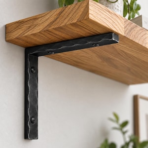 May include: A thick, light brown wooden shelf board is supported by a black, L-shaped metal bracket. The bracket features a hammered, textured surface and is secured to the wall and the underside of the shelf with visible black screws.