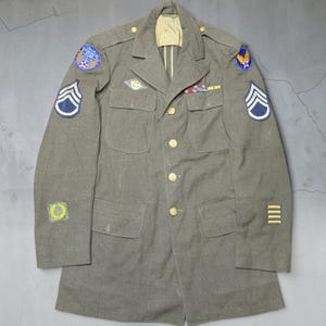 May include: An olive drab military jacket featuring a notched collar, four front pockets, and gold-toned buttons. The sleeves and chest display various embroidered patches, including a 20th Air Force emblem, rank chevrons, and service bars.