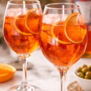 Può includere: Due candele in calici di vetro trasparente riempite con un liquido arancione vibrante, cubetti di ghiaccio e fettine sottili di arancia. Ogni candela presenta uno stoppino bianco centrato in alto, imitando l'aspetto di un cocktail.