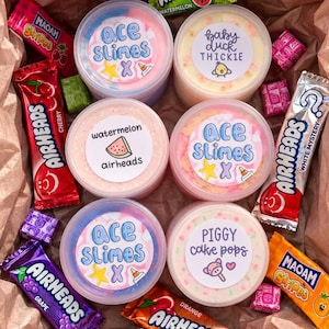Puede incluir: Seis recipientes redondos de slime de colores con etiquetas personalizadas que incluyen Ace Slimes, Baby Duck Thickie, Watermelon Airheads y Piggy Cake Pops, rodeados de varias barras de dulces Airheads y Maoam envueltas en una caja de papel.