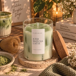 May include: A clear glass candle with a white label reading HOMEMADE ICED MATCHA LATTE 7 OZ/200 G. The candle contains pale green wax cubes resembling ice, a single white wick, and is accompanied by a round, textured cork lid placed to the side.