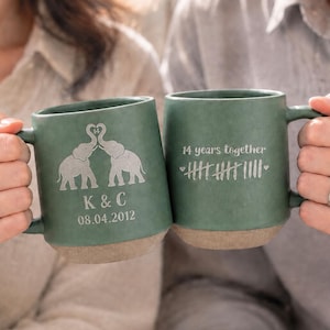 May include: Two sage green ceramic mugs with a matte finish and a raw, unglazed base. One mug features two elephants forming a heart with their trunks, the initials K & C, and the date 08.04.2012. The other mug displays 14 years together with tally marks.