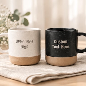 Peut inclure: Deux tasses en céramique avec une finition mouchetée, l'une blanche et l'autre noire, chacune dotée d'une base beige. La tasse blanche porte l'inscription "Your Own Logo" et la tasse noire "Custom Text Here" dans une police sombre en creux.