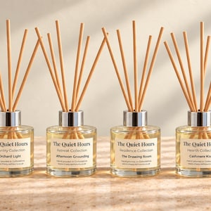 May include: Six clear glass reed diffusers with silver collars and wooden reeds. Each bottle features a white label with the text The Quiet Hours, a specific collection name, and a scent name including Sea Glass, Orchard Light, and Midnight Library.