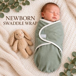 May include: A sage green newborn swaddle wrap with white trim and a hook-and-loop closure, featuring a sleeping infant wrapped inside. The text NEWBORN SWADDLE WRAP is printed in brown. A small beige plush bunny toy sits next to the swaddled infant.