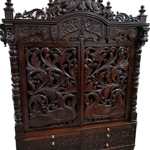 Può includere: Un mobile in legno marrone scuro con intricati motivi floreali e disegni di pavoni intagliati a mano sulle due ante principali. Il pezzo include due cassetti inferiori con piccoli pomelli rotondi e un frontone superiore intagliato.